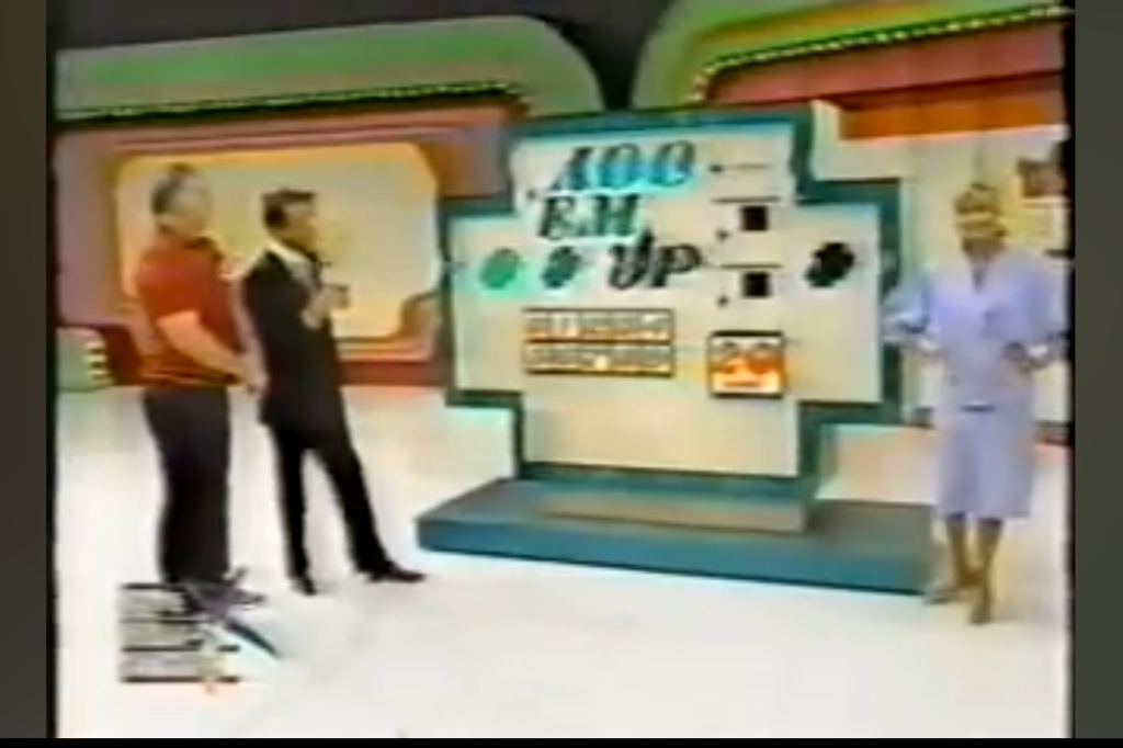 A scene from the game 'Add 'Em Up' featuring a host interacting with contestants in front of a colorful game set.