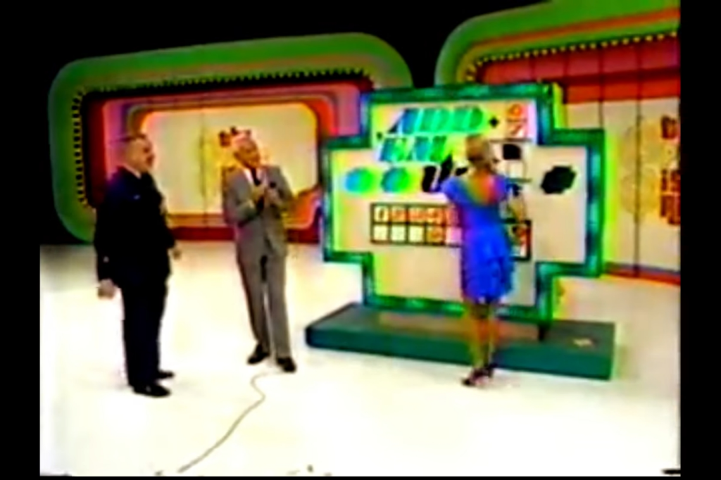 A colorful set from the game 'Add 'Em Up' on a game show, featuring a host and two contestants. The host is engaging with the audience while one contestant stands ready to play.