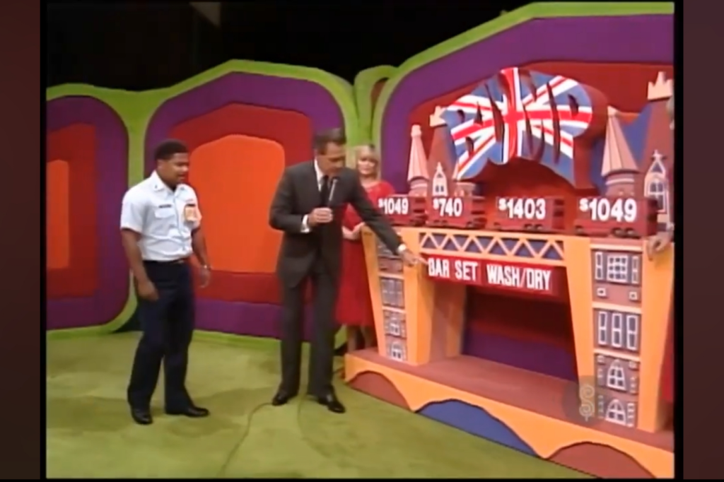 A game show set on The Price is Right featuring contestants and a host beside colorful display boards with prices and items labeled, including 'Bar Set' and 'Wash/Dry'.