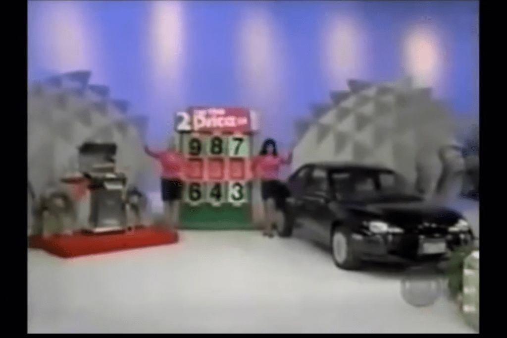 A game show set featuring a large prize display and two models. One model is standing next to a black car, while the other is by a prize showcase. The background is colorful with a light blue hue.