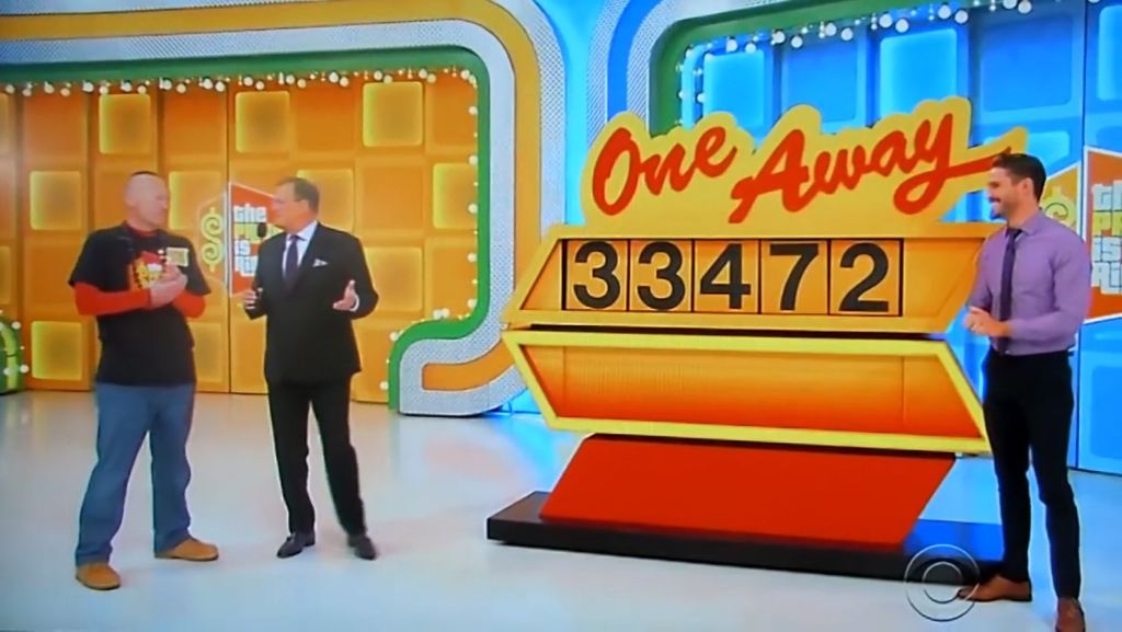 A contestant speaks with the host on the One Away game show set, with a display showing the number 33472.