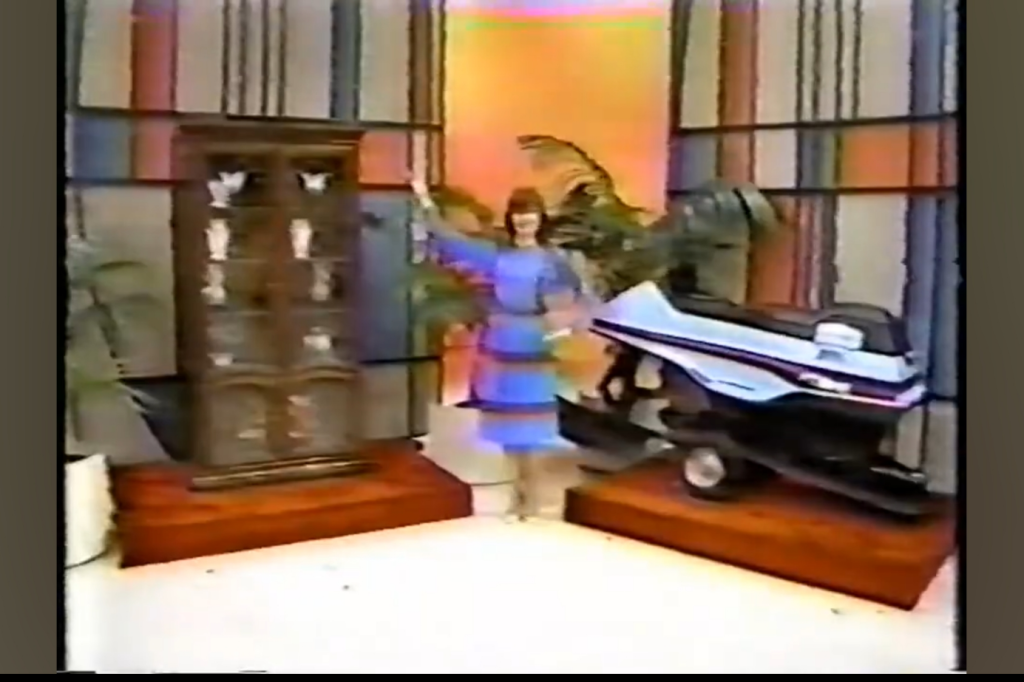 Holly Hallstrom in a blue dress stands between a glass front cabinet displaying decorative items and a snowmobile, set against a colorful backdrop.