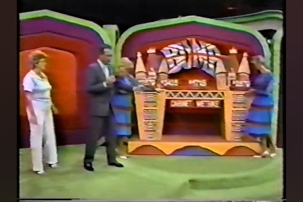 The set of The Price is Right featuring a stage with decorative towers, with Bob Barker, a contestant, and two models, one on each side of the stage. The host is speaking, while the models are engaged with the set.