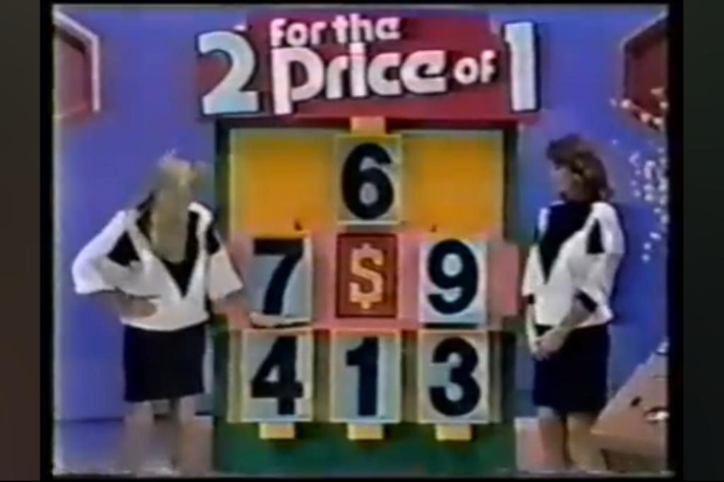 Two women standing next to a colorful game board displaying the text '2 for the Price of 1' and numbers 6, 7, 9, and 413.