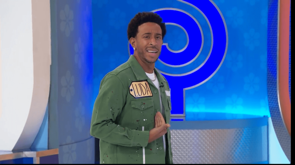 Ludacris wearing a green jacket with a name tag stands on the set of The Price is Right, smiling and gesturing.