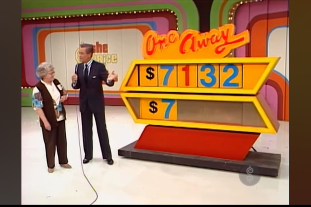 Contestant standing with host Bob Barker, discussing the game 'One Away' with the price ,132 displayed on the game board, and a blue 7 underneath matching.