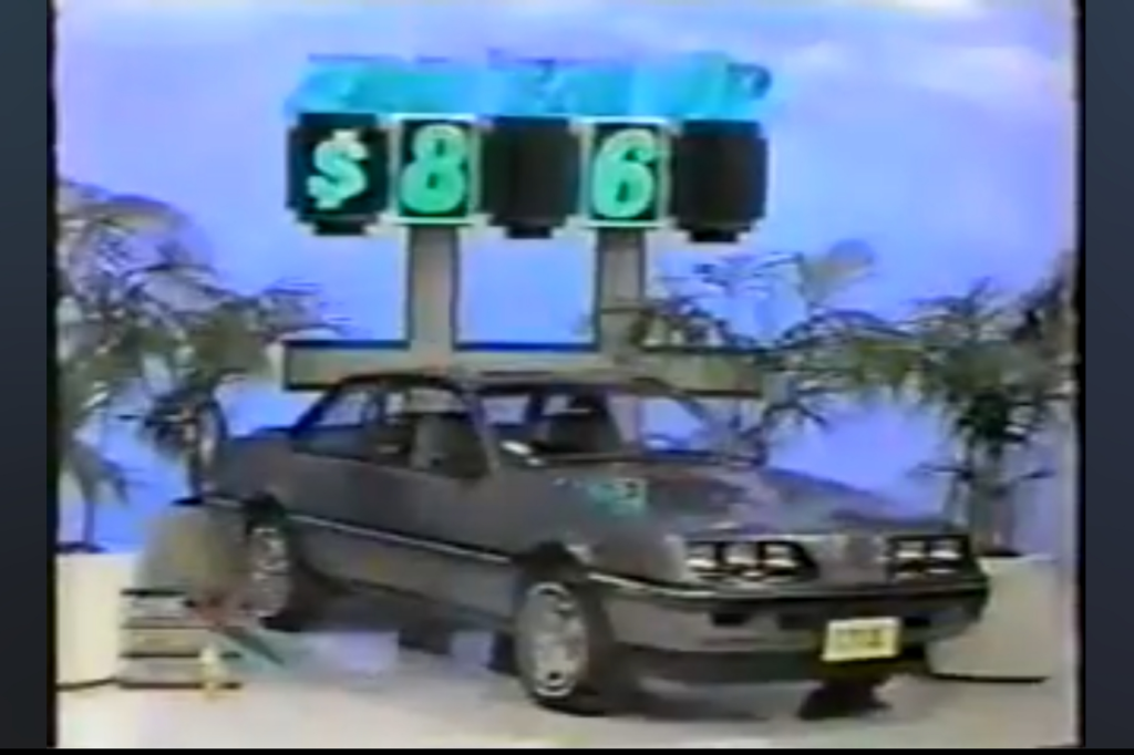 A gray Pontiac Sunbird Coupe displayed with a large game board showing the partial price of __6 for a game called 'Add 'Em Up.'