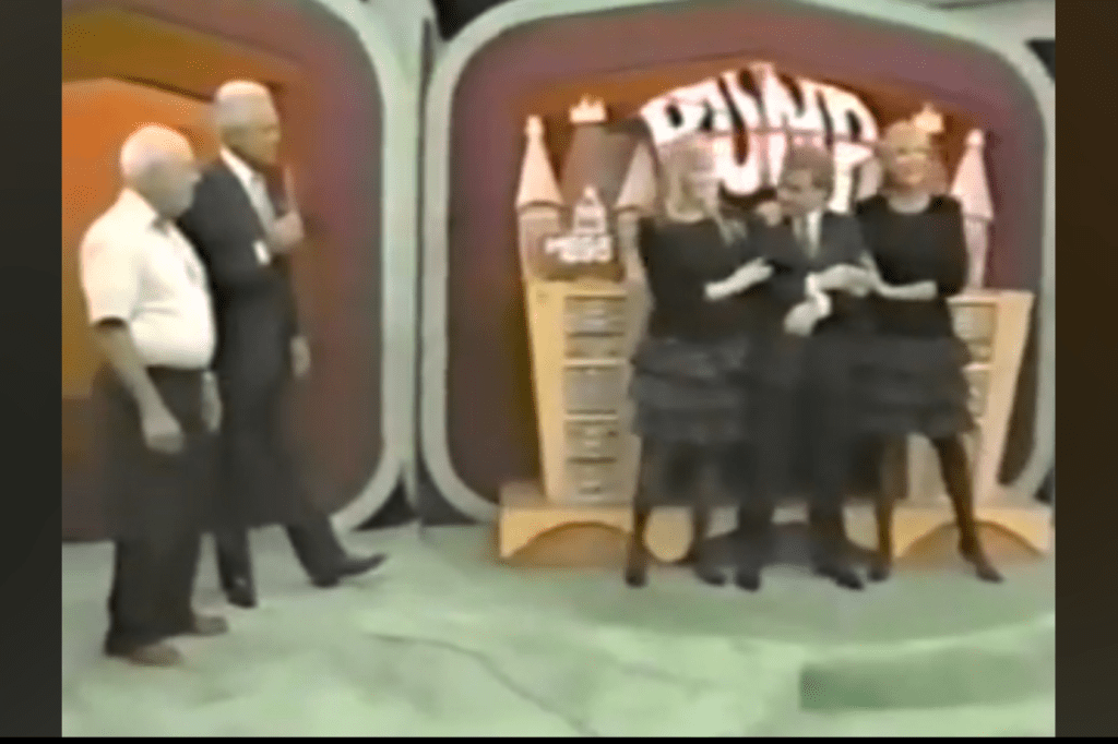 A vintage television game show scene featuring four people: two men standing on the left and two women in black dresses on the right, one of whom is holding a man in a suit. The background displays a colorful game show set with a sign reading 'Wheel of Fortune'.