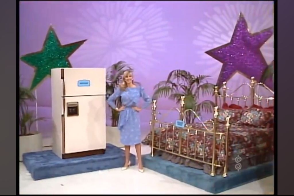 A contestant stands next to a large refrigerator and a decorative bed set, both showcased in a game show setting with colorful star decorations in the background.