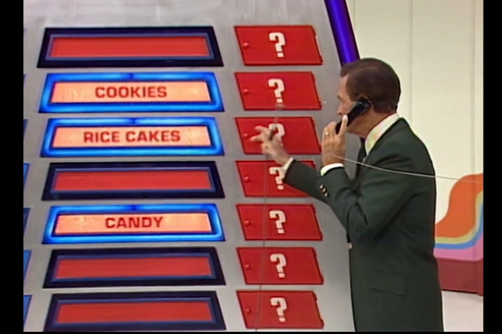 A game show host on a call holds a microphone while pointing at three visible grocery items labeled 'Cookies', 'Rice Cakes', and 'Candy' on a game board with red and blue panels.
