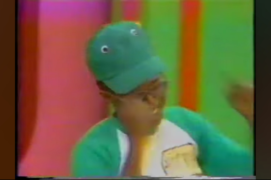 A contestant wearing a green and white outfit and a green hat with eyes, celebrating or dancing, in a colorful studio backdrop.
