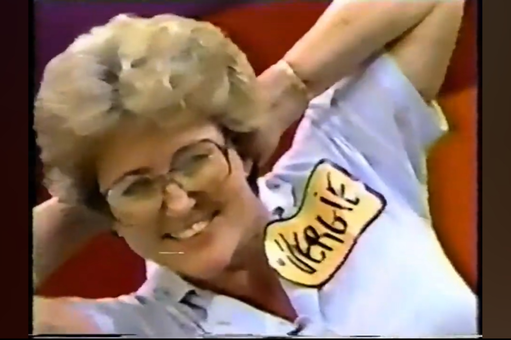 A smiling woman with glasses and curly hair wears a shirt with a name tag reading 'Vergie', posing playfully.