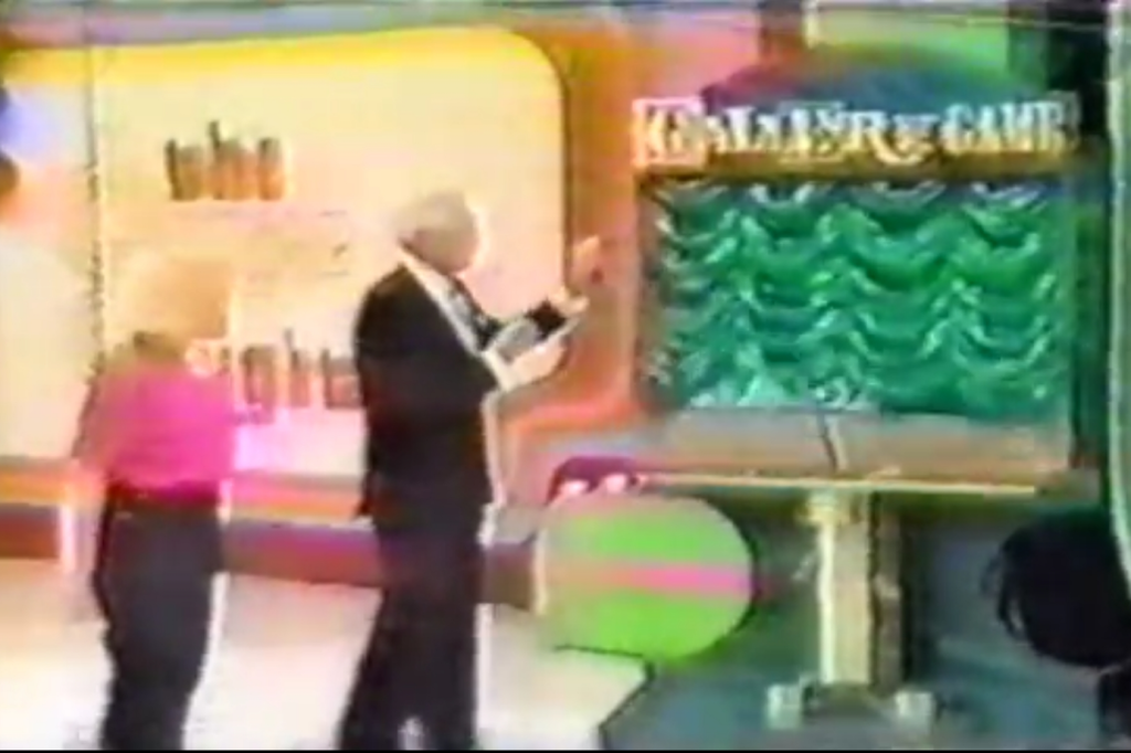 A game show set featuring a host in a black suit and a contestant in a pink top, standing next to a green game board with the title 'GALLERY GAME' displayed.