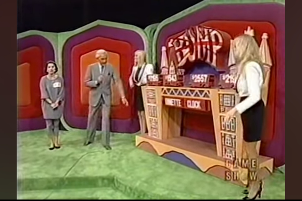 A scene from The Price is Right featuring a host speaking to two female contestants. One contestant is standing to the left, while the other is next to a game board displaying various numbers and the word 'BUMP.' The set has colorful backgrounds with green carpet.