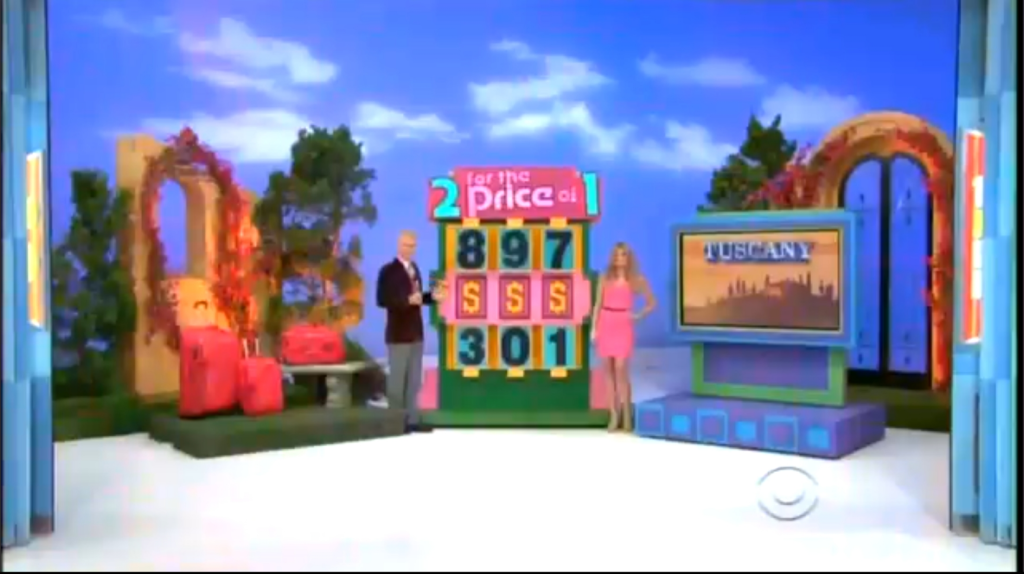 A game show set featuring announcer George Gray and a model beside a game board labeled '2 for the Price of 1' with numbers and a dollar sign. In the background, there is a sign reading 'Tuscany' next to decorative arches and luggage.
