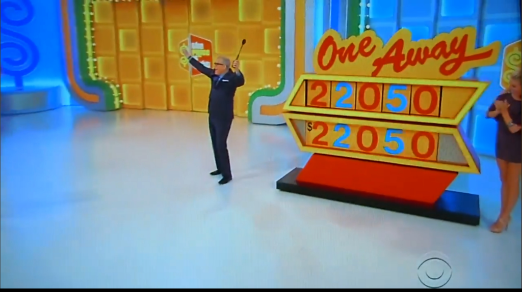 A game show host stands on stage with a pointer, presenting the One Away game board displaying the price '22050'. A female model stands beside him, applauding.