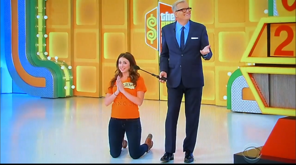 Contestant kneeling on stage with hands clasped, looking hopeful while host stands beside her, set against a colorful game show backdrop.