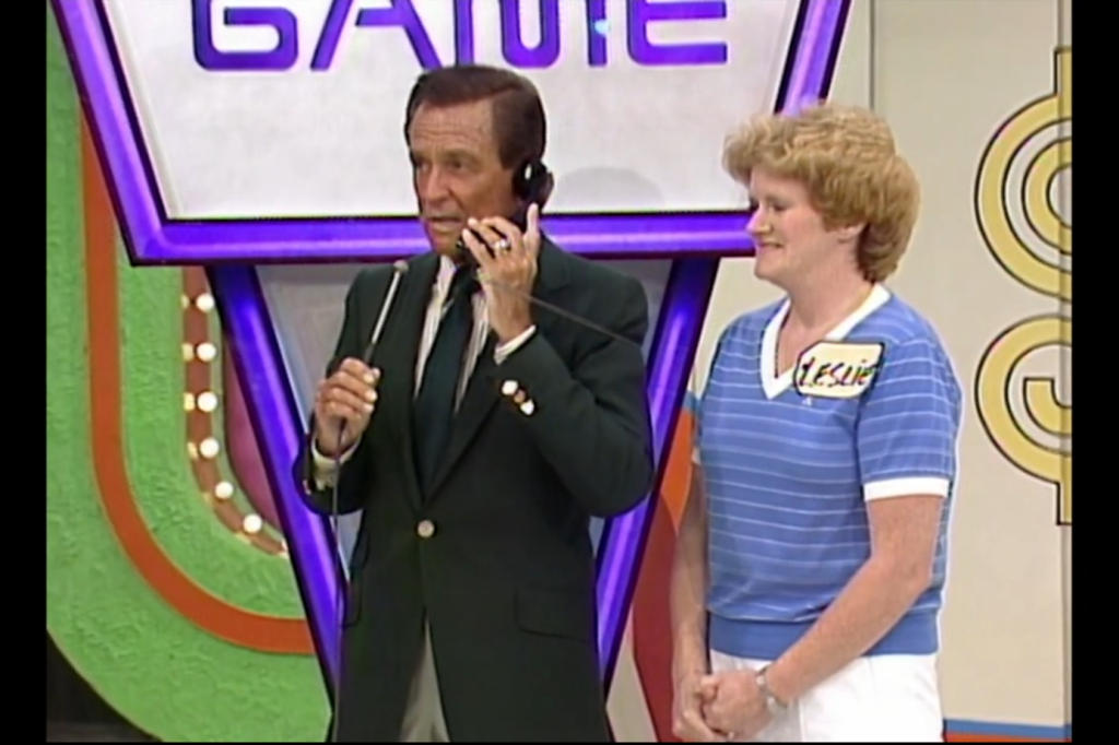 A game show host speaking on the phone while a contestant stands beside him, both set against a colorful background displaying the words 'PHONE HOME GAME'.