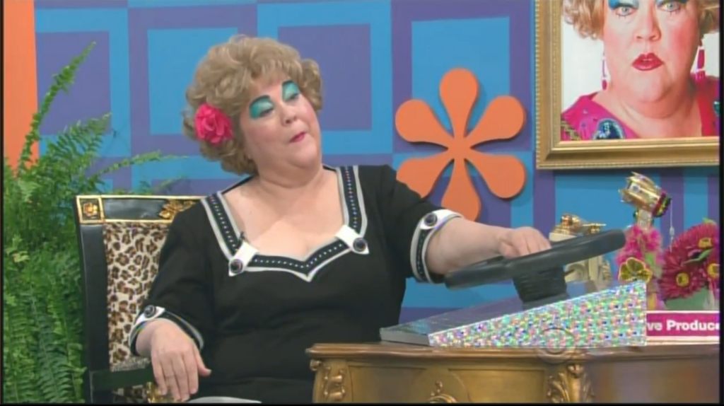 Mimi Bobeck, a woman with styled hair and bright makeup, wearing a black and white outfit, sits at a game show console holding a steering wheel, with a colorful backdrop featuring a floral arrangement and a framed photo.