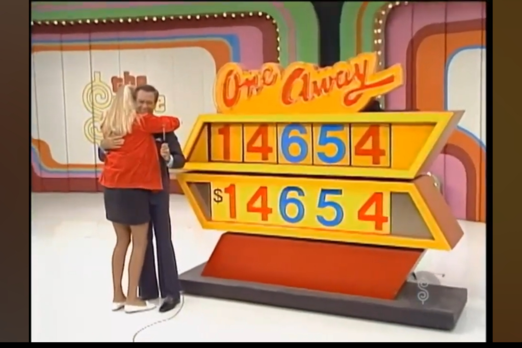 A contestant hugs the host on stage during the game 'One Away', displaying a pricing board with the numbers 14,654.
