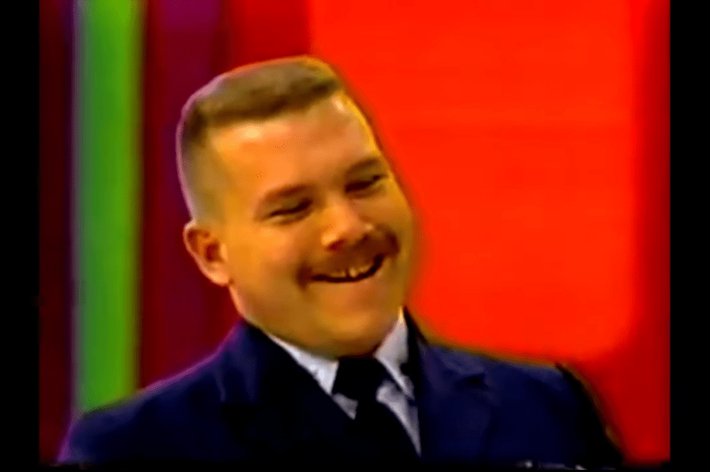 A smiling contestant on a game show, dressed in a dark suit, with a colorful backdrop.