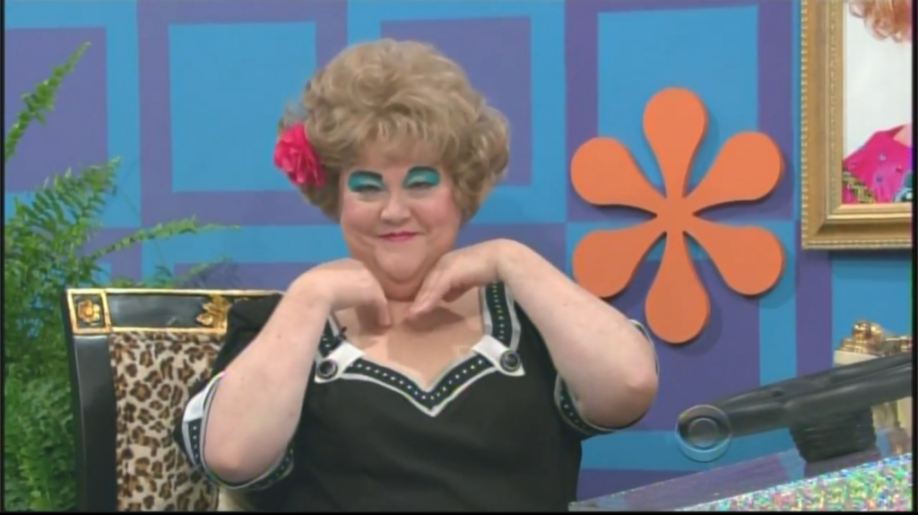 Mimi Bobeck from the Drew Carey Show with a large hairstyle and bright makeup poses playfully with her hands under her chin, set against a colorful background.
