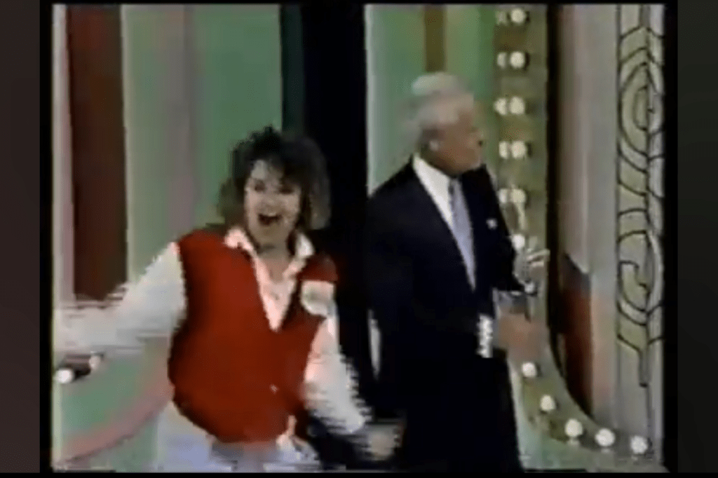 A person in a red vest cheerfully gestures while standing next to Bob Barker, both in front of a colorful backdrop with lights on the set of The Price is Right.