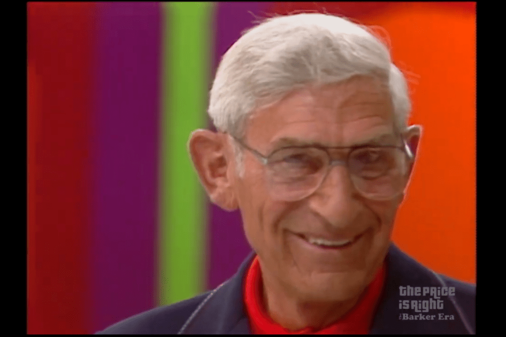 A smiling older man wearing glasses and a dark jacket, with a colorful background, likely from a game show.