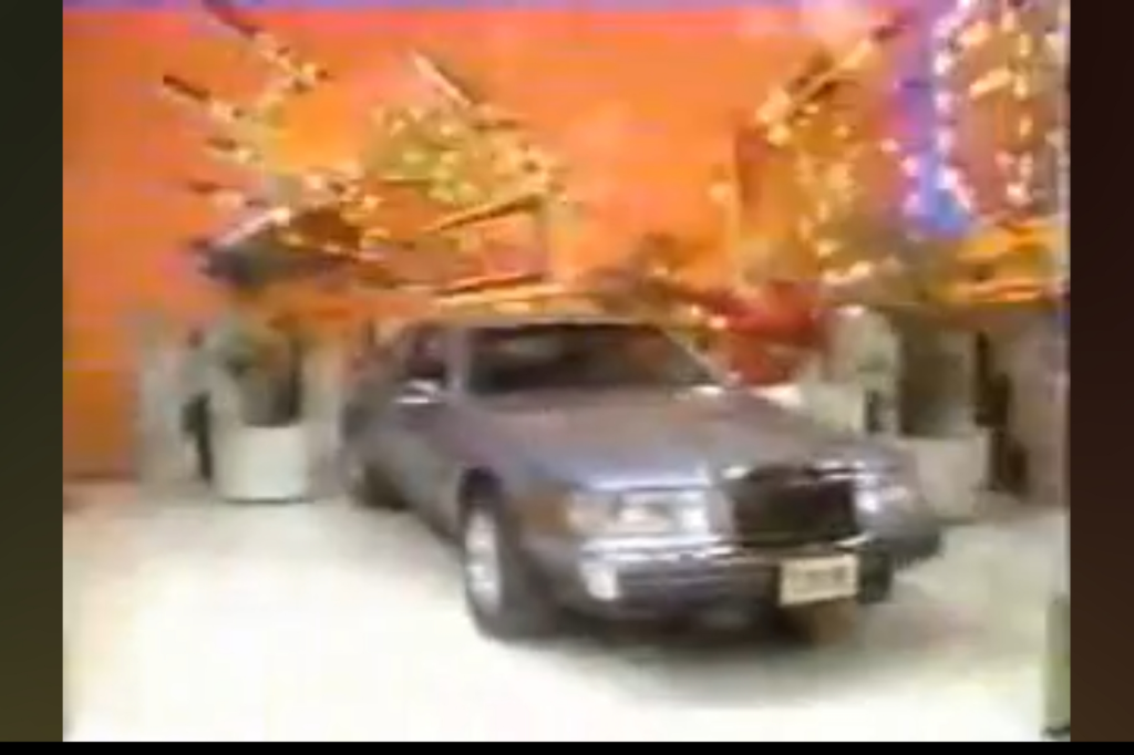 A silver car showcased on a brightly colored game show set, surrounded by cheering audience members and decorative lights.