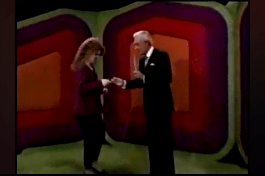 A man in a black suit stands on the set of The Price is Right, handing a 0 bill to a woman in a maroon outfit.