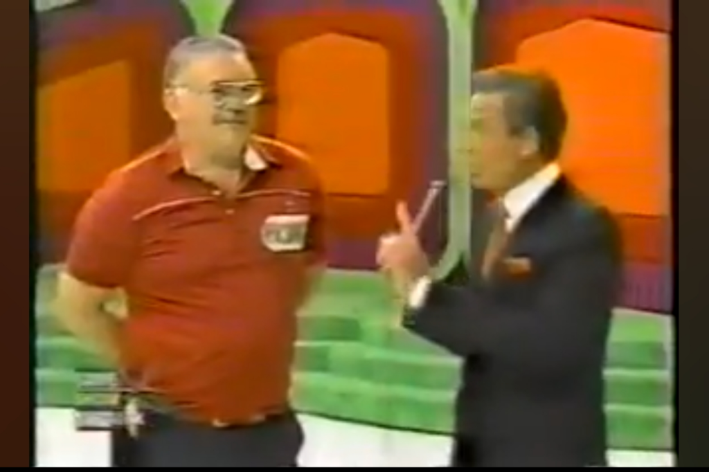 A game show contestant wearing a red shirt stands with his hands behind his back, while the host gestures toward him with a pointer, set against a colorful game show backdrop.
