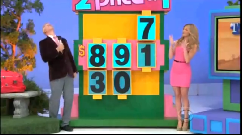 A game show scene featuring George Gray in a dark blazer and a pink tie, expressing excitement while looking upward, alongside a female model dressed in a pink dress, standing next to a large colorful game board displaying the numbers 89130 and a highlighted 7.