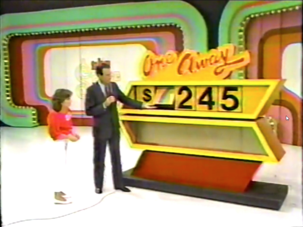 A game show host is explaining the One Away game to a young female contestant, with a large board displaying an incorrect car price of ,245. The host demonstrates that the first number could rotate up to a blue 7.
