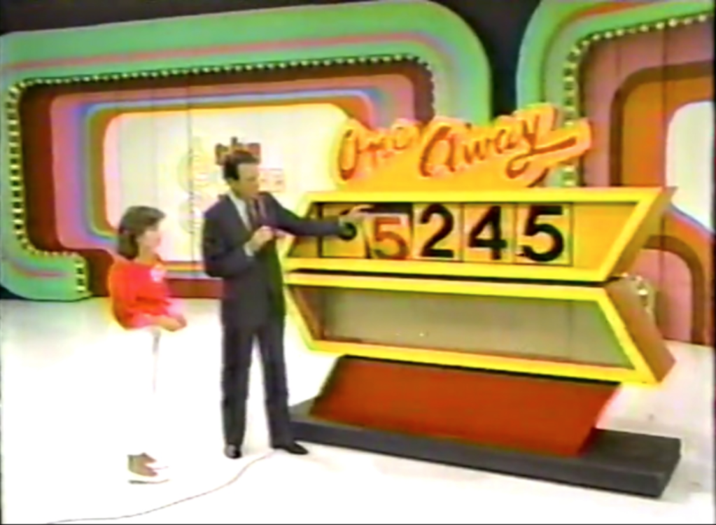 A game host explains the pricing game 'One Away' while a young contestant looks on, with a display board showing the incorrect price of ,245.