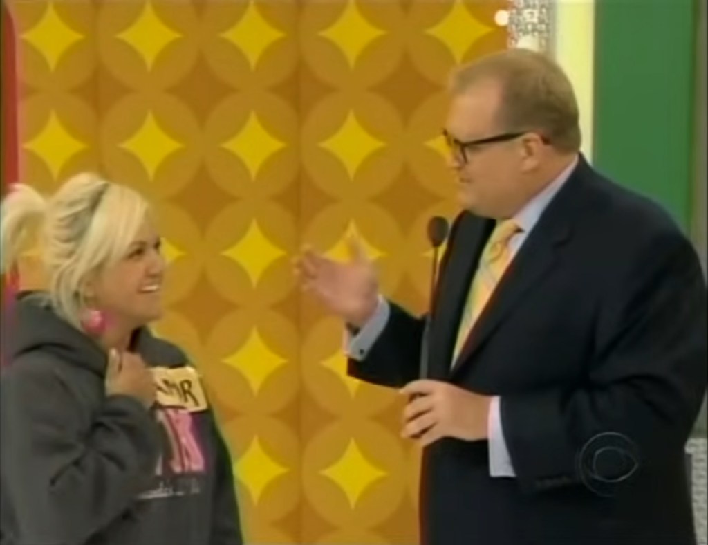 A contestant smiles while speaking to host Drew Carey on stage during a game show, with a colorful backdrop featuring a yellow and orange design.