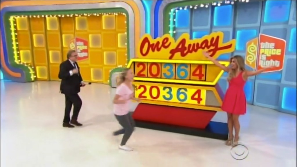 A contestant joyfully plays the One Away game on The Price is Right, standing near a large game display with numbers, as the host looks on.