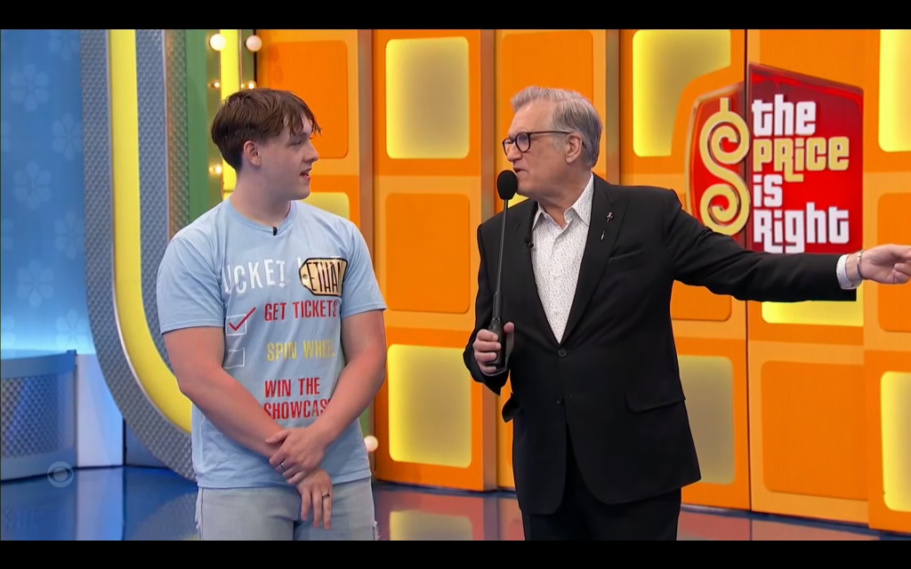 A contestant named Ethan stands on stage during a game segment of 'The Price is Right', speaking with the host, who gestures while holding a microphone.
