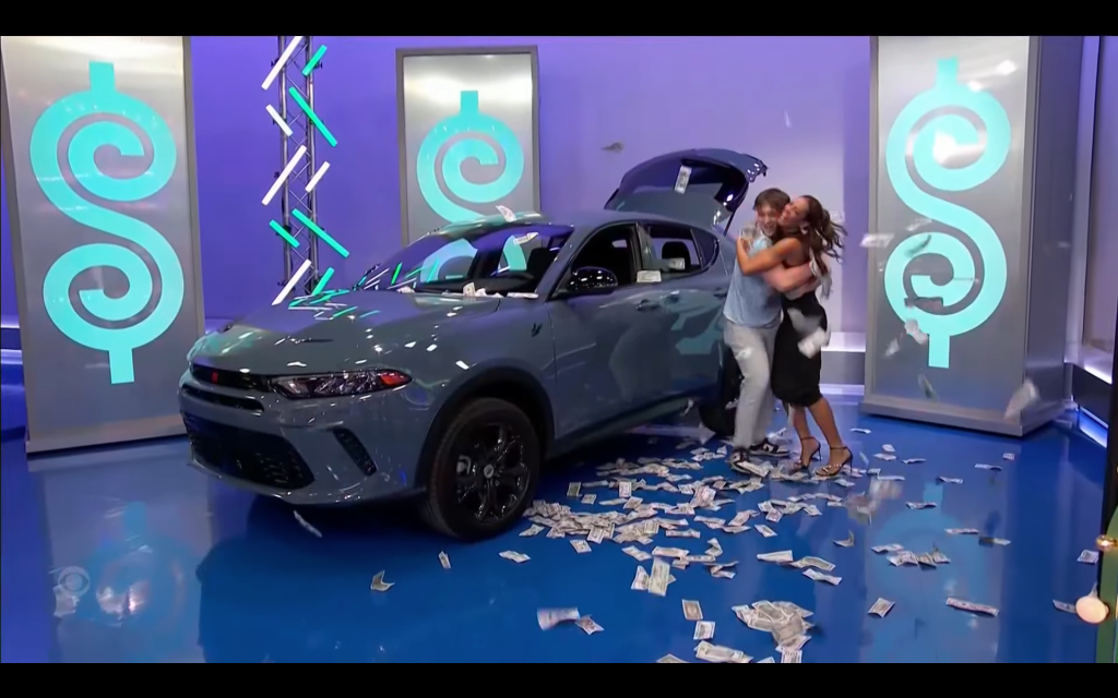 A couple joyfully celebrates in front of a new gray car, surrounded by confetti and fake money, during a game show.