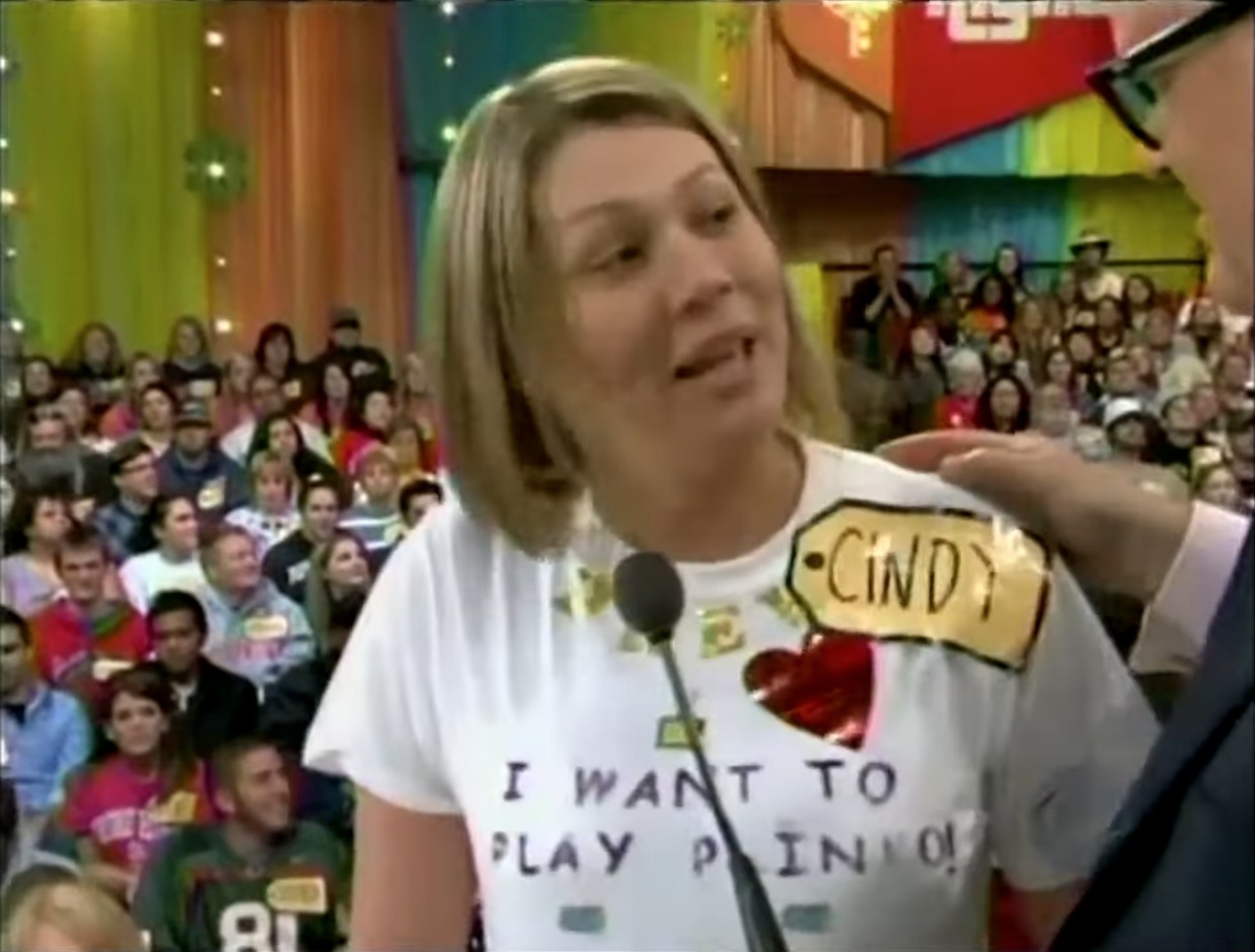 A contestant named Cindy, wearing a shirt that says 'I WANT TO PLAY PLINKO', interacts with the host on stage during a game show, with an excited audience in the background.