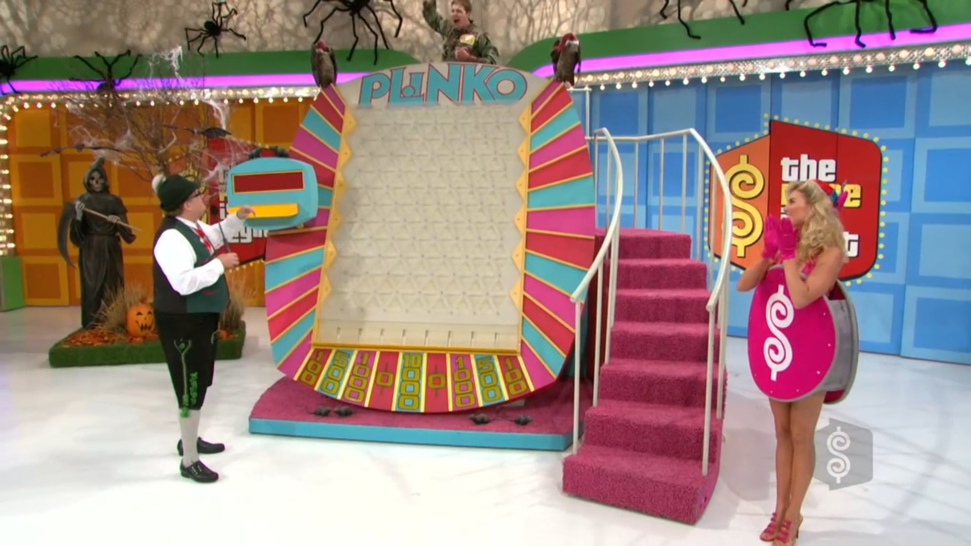 A colorful Plinko board set up for a game on The Price is Right, featuring the word 'PLINKO' at the top, with a contestant holding a Plinko chip and a model in a money-themed costume standing beside him.