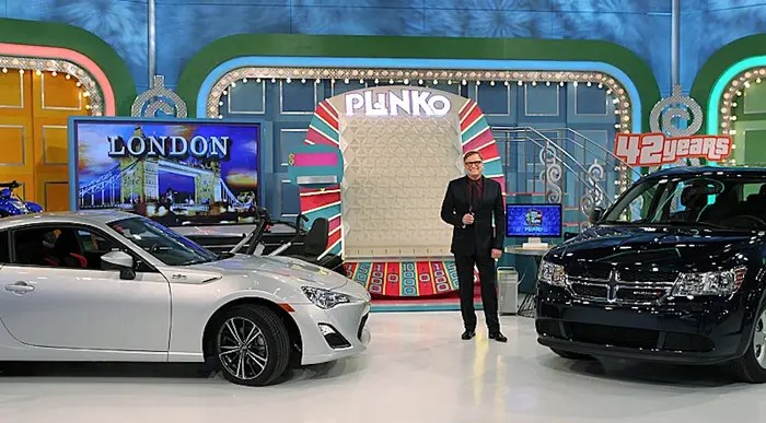 A host stands next to a large Plinko board on the set of The Price is Right, with two cars displayed on either side and a screen showing 'LONDON' in the background.