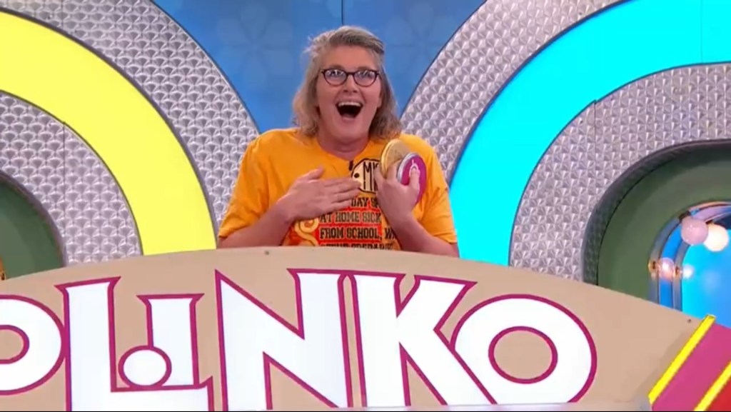 A contestant with glasses and a yellow shirt smiles excitedly while holding a Plinko chip, standing in front of the Plinko game board.