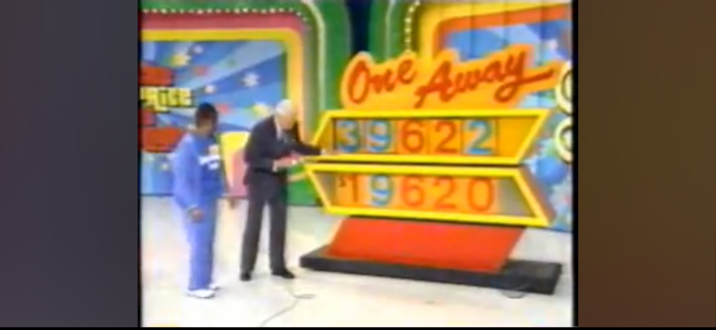 A contestant and a host interact with the game 'One Away' on stage, showcasing a colorful board displaying two sets of numbers.