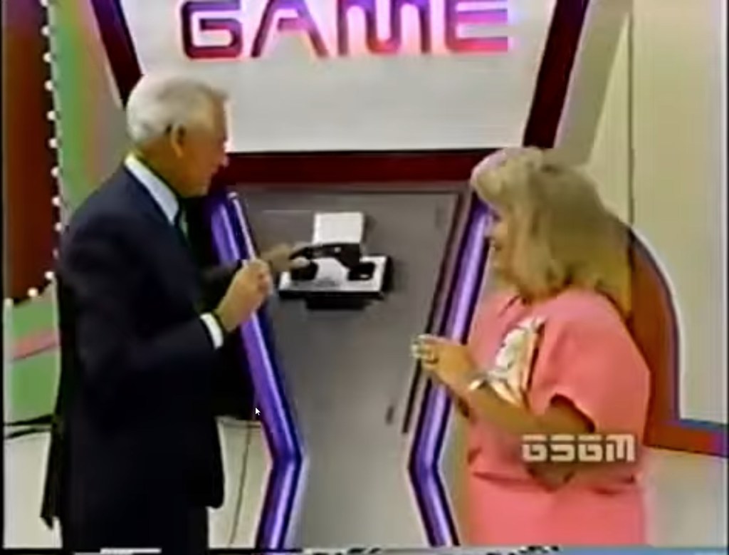 A game show host demonstrates the phone used in The Phone Home Game on The Price is Right, with a female contestant looking on excitedly.