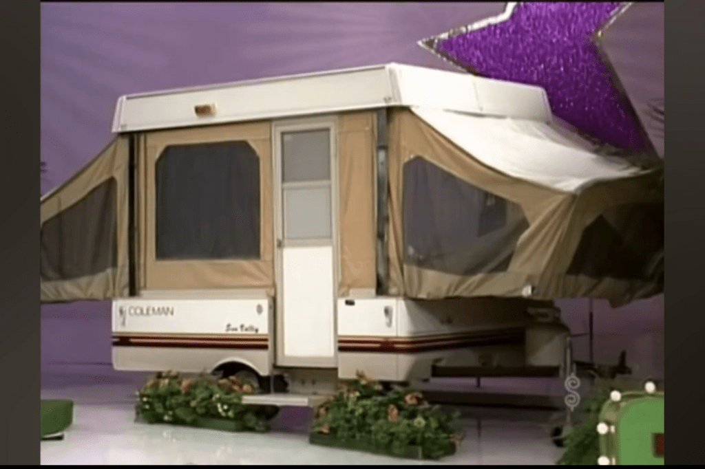 A Coleman pop-up camper trailer displayed as a prize on a game show set, adorned with flowers and a purple star backdrop.