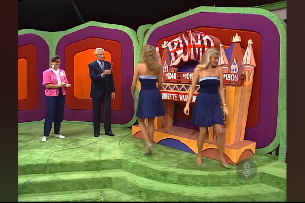 Two women in blue dresses walk in front of a colorful game show backdrop with the word 'BUMP.' A man in a dark suit holds a microphone, while a woman in a pink shirt observes.