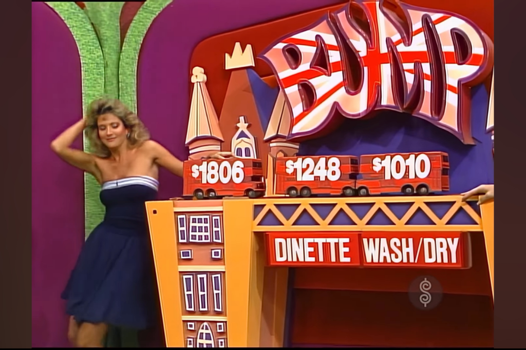 Kyle Aleter in a blue dress is playfully interacting with a game setup displaying prices of items on a stage, with a colorful backdrop and the word 'BUMP' prominently featured.