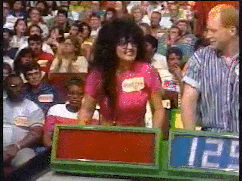 A woman with curly hair wearing a pink shirt bounces in place while standing next to a man in a striped shirt on the set of The Price is Right, with a cheering audience in the background.