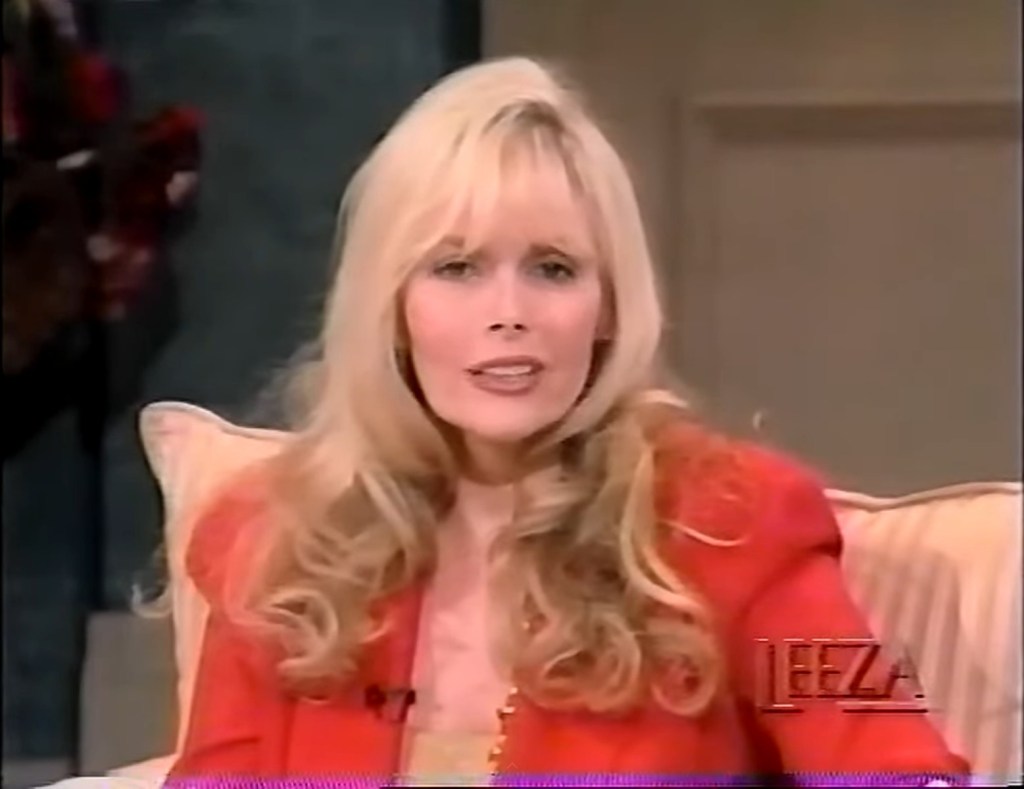 Dian Parkinson with long blonde hair wearing a red jacket speaks during a television interview. She is seated on a sofa, appearing engaged and expressive.