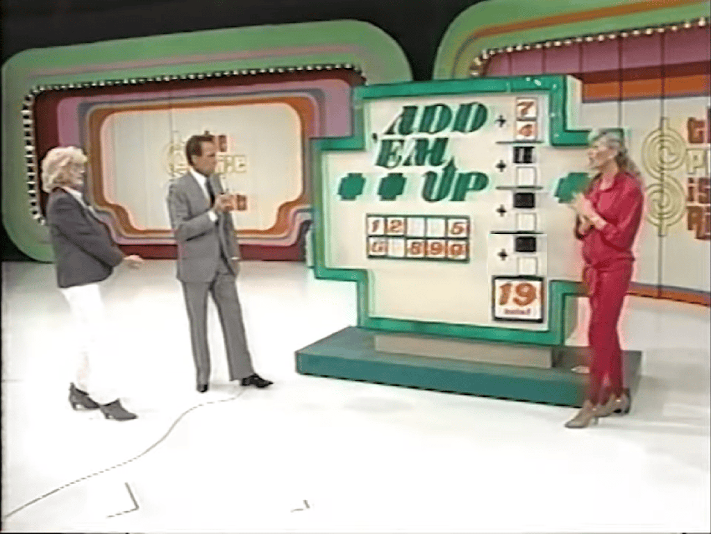 A game show set for 'Add 'Em Up' featuring a large board with numbers and a colorful backdrop, while two contestants and a host engage in the game.